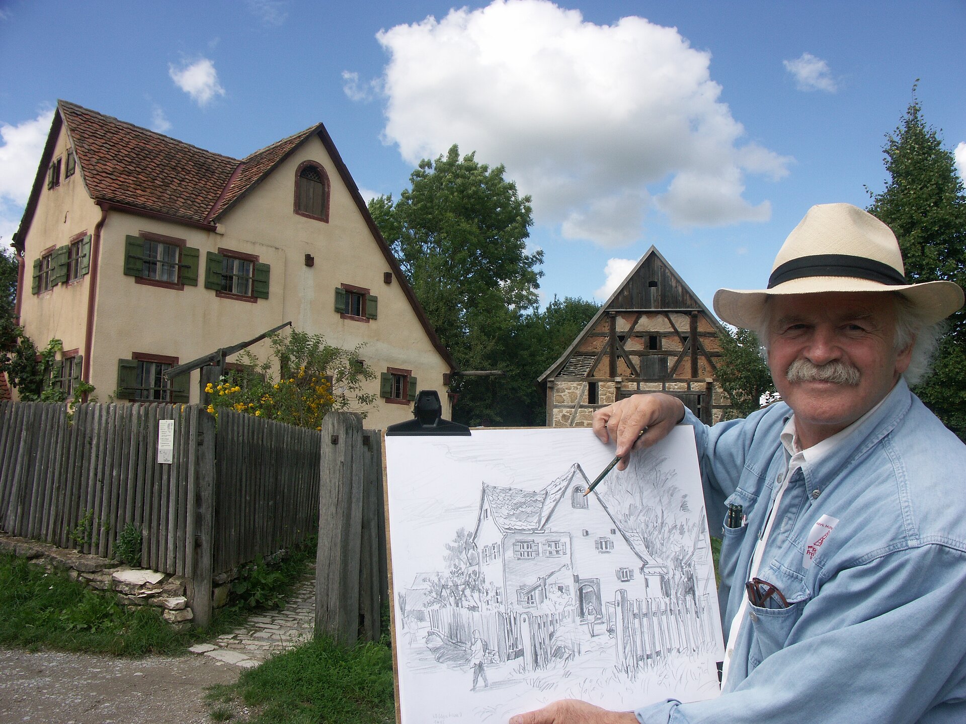 Künstler im Vordergrund präsentiert sein gemaltes Werk von einem Bauernhaus im Freilandmuseum